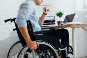 A man in a wheelchair working on a laptop behind a desk.